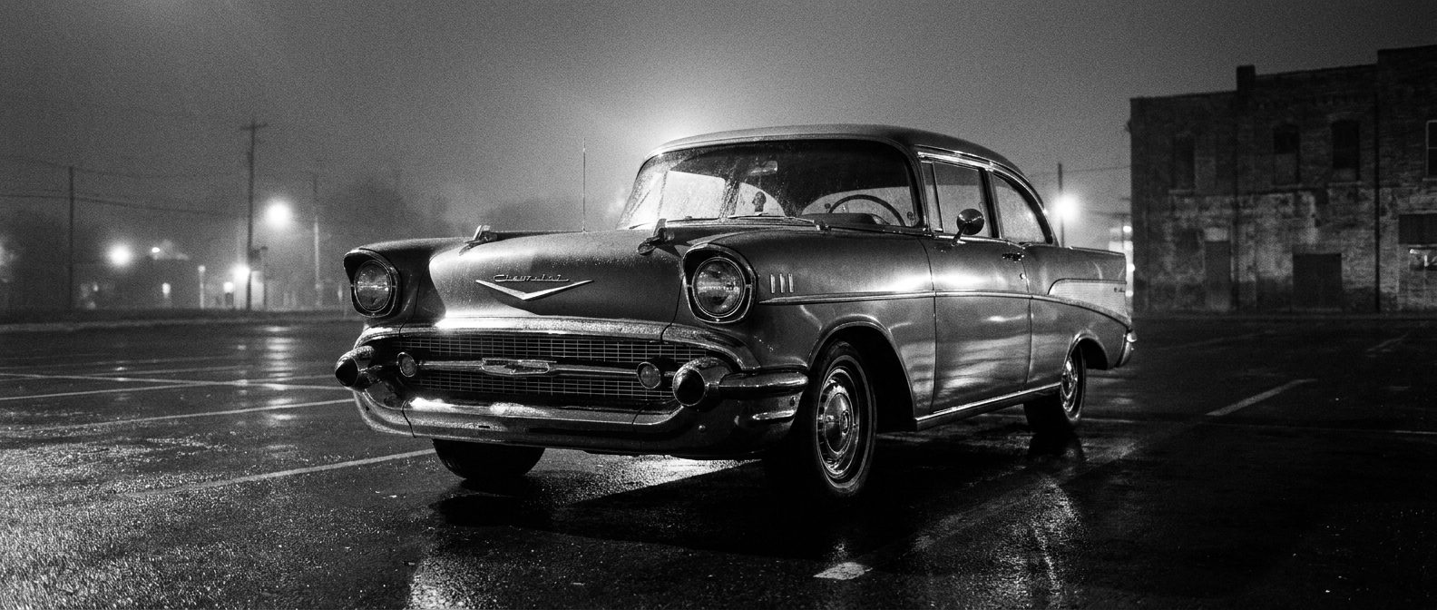 Classic chrome-grille car on wet asphalt at night, black and white
